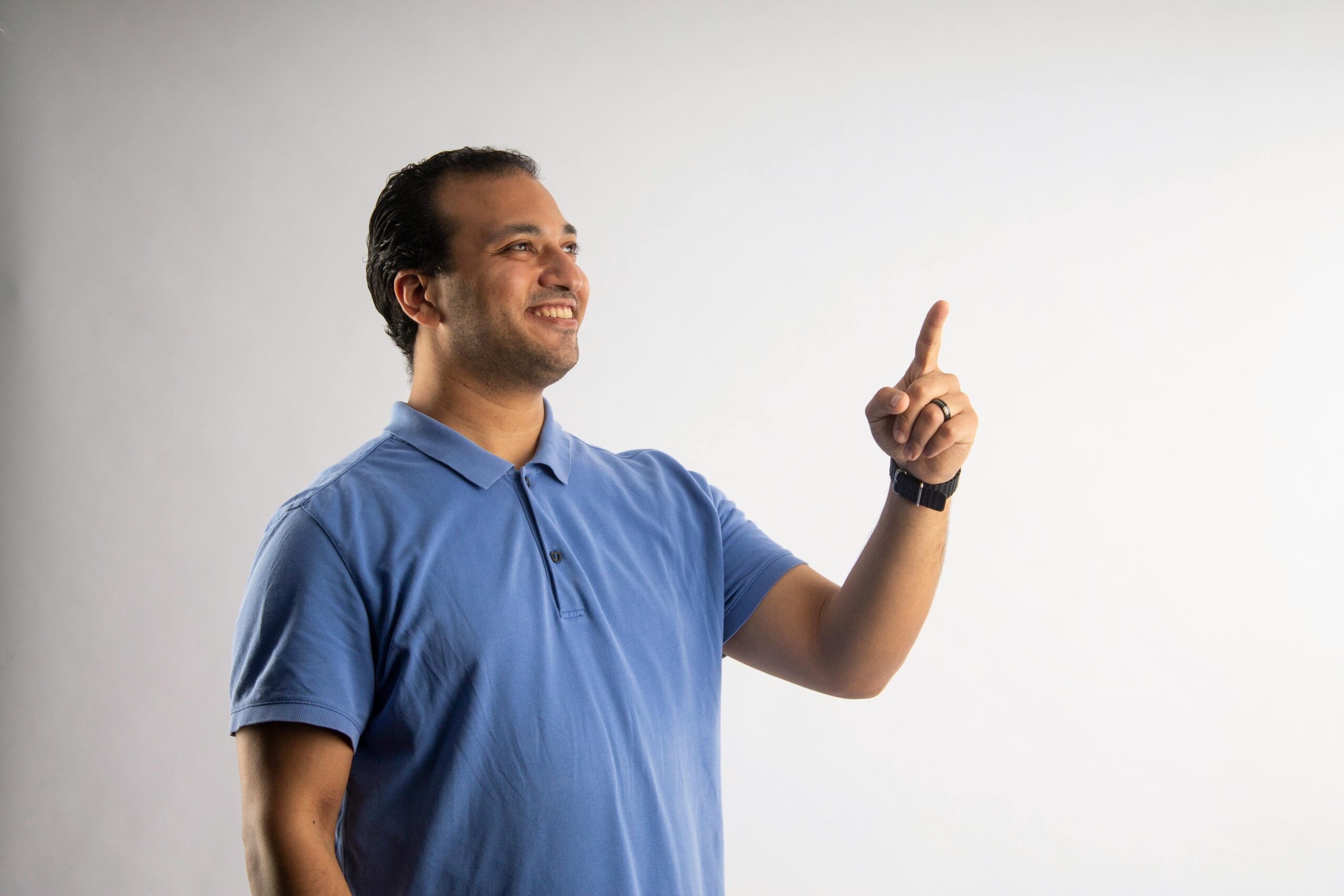 A confident man in a blue shirt points upward, smiling against a plain white background.